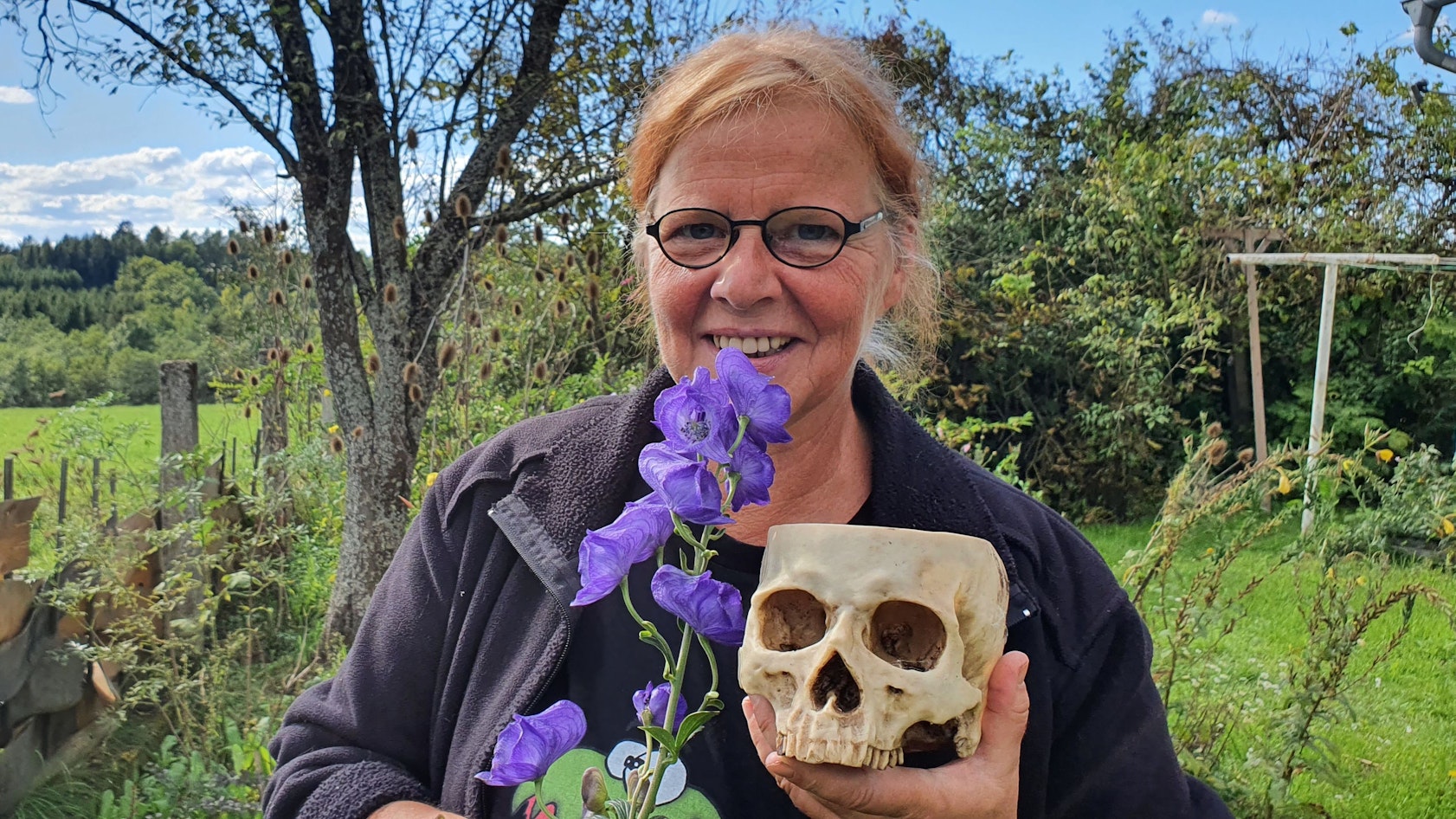 Eine Frau mit schwarzer Brille und Jacke steht in ihrem Garten und hält eine lila geblümte Pflanze und einen Totenkopf in den Händen.