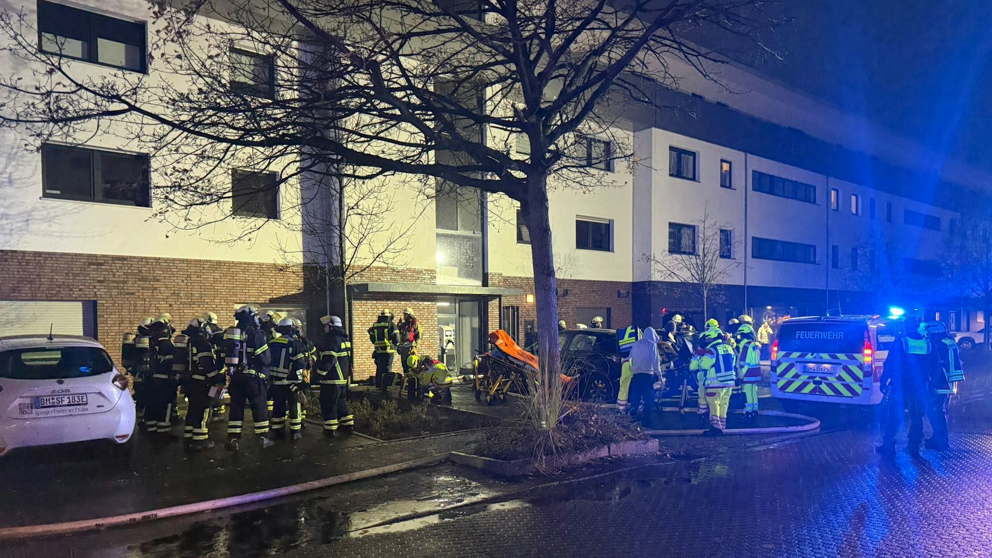 Das Foto zeigt Feuerwehrleute und ein Einsatzfahrzeug mit Blaulicht vor dem Mehrfamilienhaus.