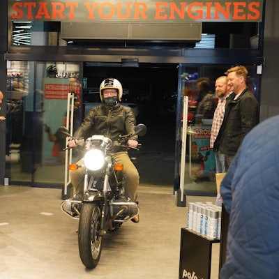 Auf dem Bild ist ein Mann zu sehen, der bei einer Eröffnungsfeier eines Geschäfts mit einem Motorrad in das Ladenlokal fährt.