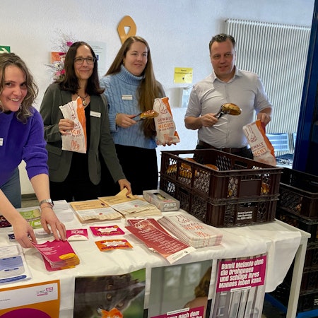 Zwei Mitarbeiterinnen der Frauenberatungsstelle, die Gleichstellungsbeauftragte und der Bürgermeister stehen hinter dem Infostand, an dem es Broschüren und Flyer gibt sowie Weckbrötchen, die in die speziellen Tüten gepackt werden.
