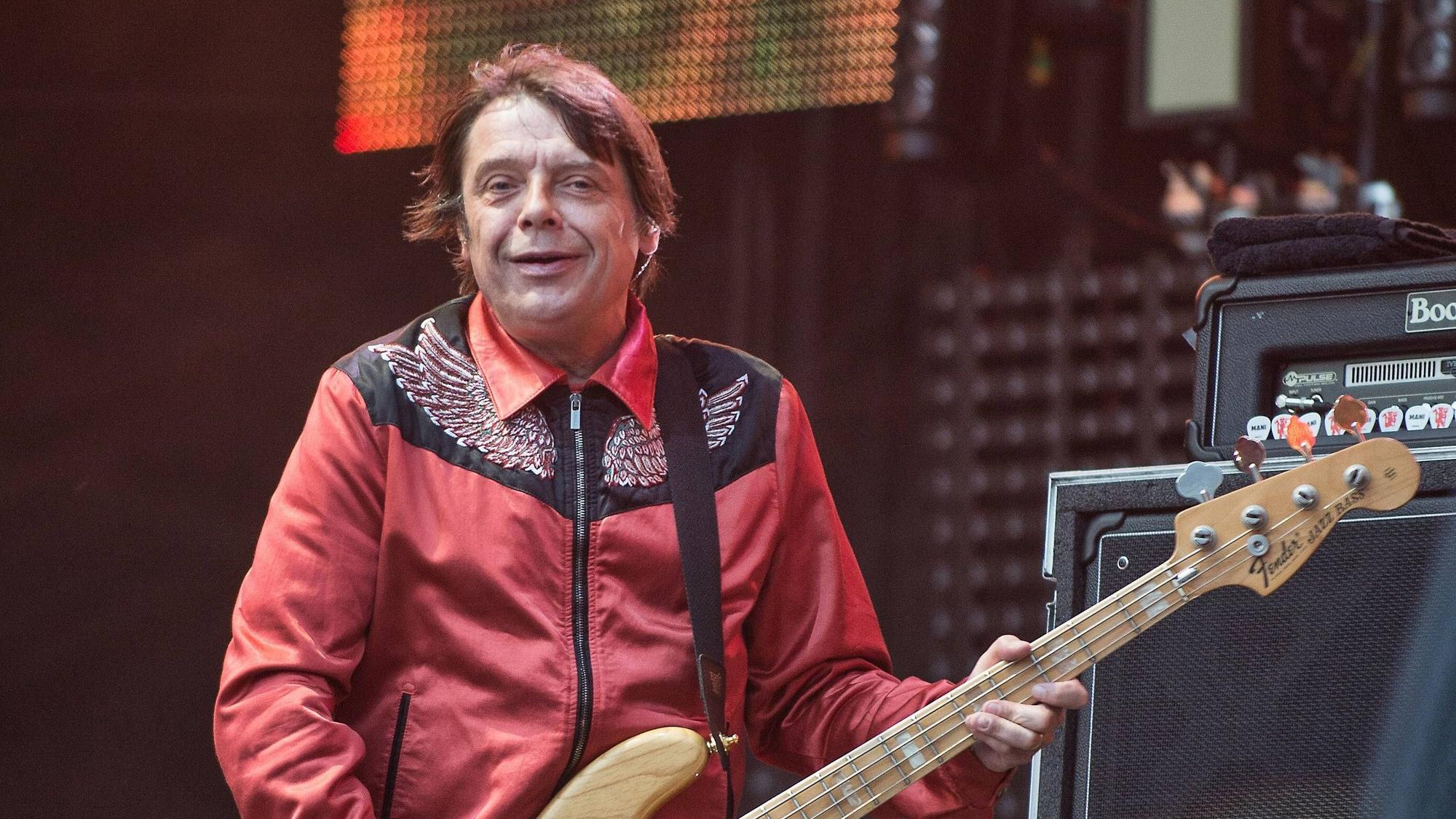 Mani von The Stone Roses spielt Bassgitarre auf der Bühne, trägt eine rote Jacke