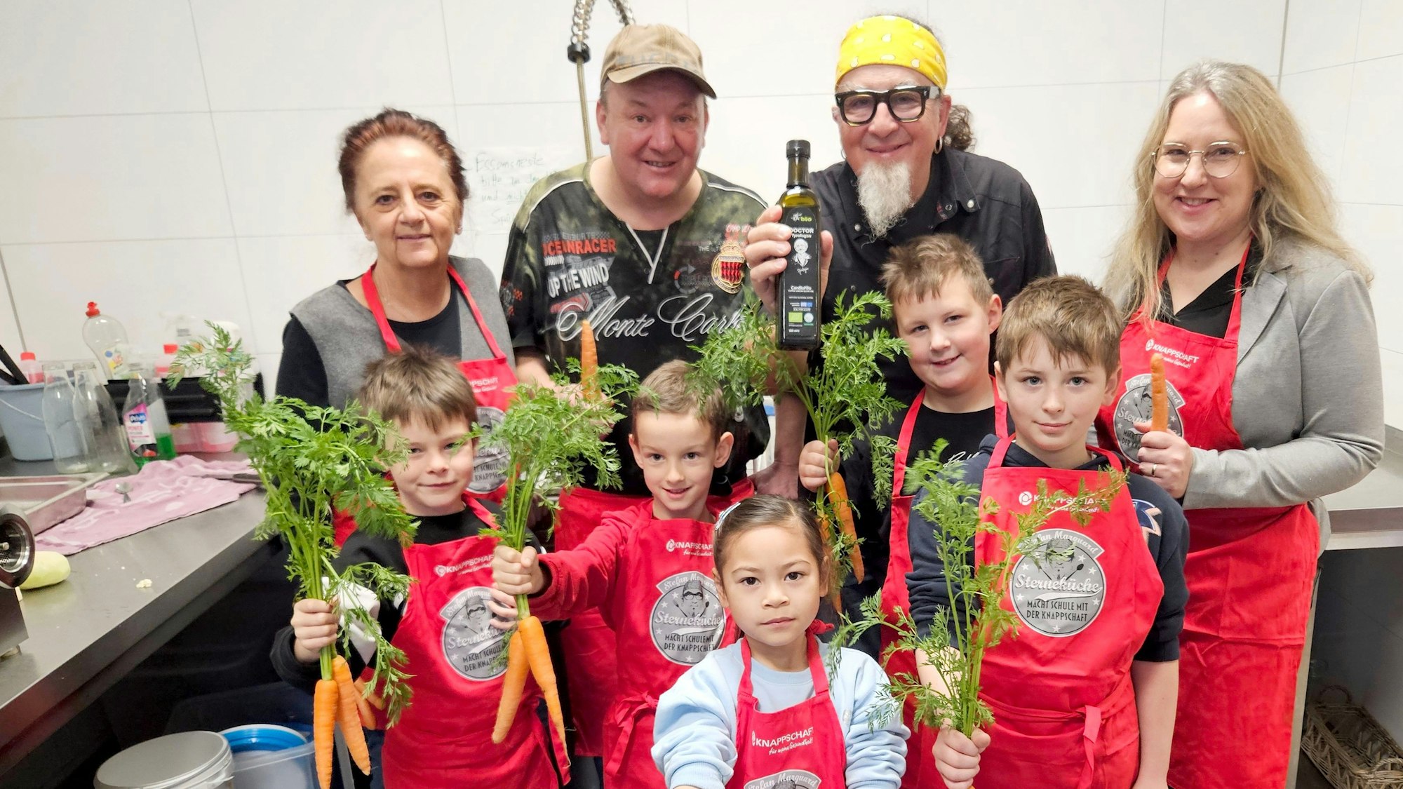 Gruppenfotos des Kochteams: Die Kinder, auch mit roten Schürzen ausgestattet, halten Mohrrüben in den Händen.
