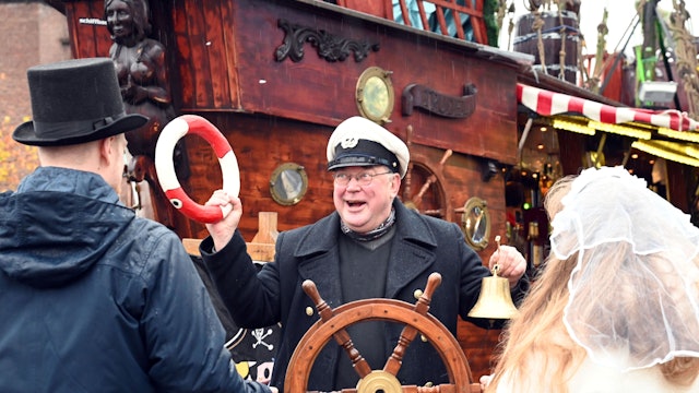 Ein Mann mit Kapitänsmütze hält einen kleinen Rettungsring und eine Glocke in den Händen. Vor ihm steht ein Paar, der Mann mit Zylinder, sie mit Schleier. Gemeinsam halten sie ein Steuerrad.