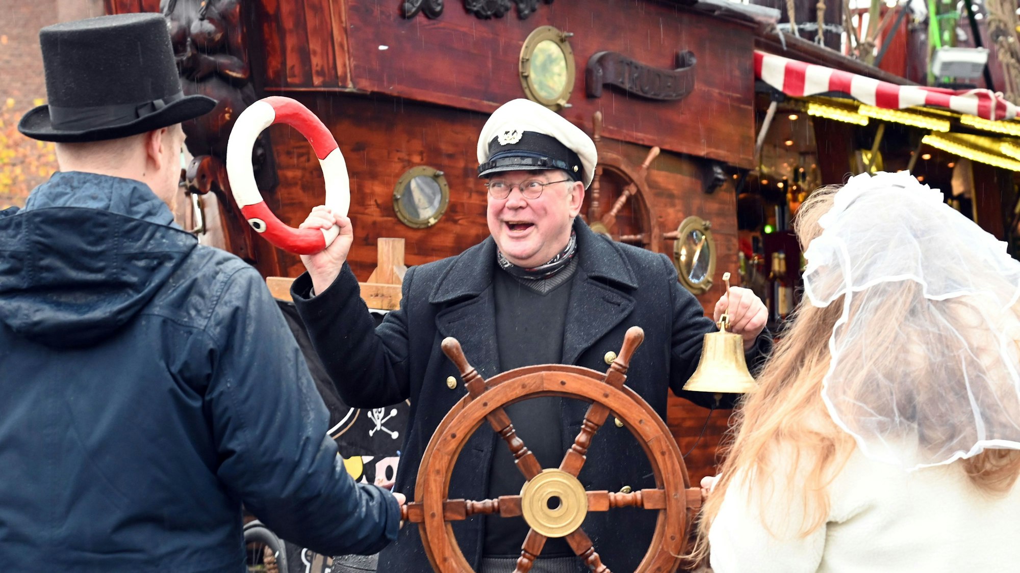 Ein Mann mit Kapitänsmütze hält einen kleinen Rettungsring und eine Glocke in den Händen. Vor ihm steht ein Paar, der Mann mit Zylinder, sie mit Schleier. Gemeinsam halten sie ein Steuerrad.