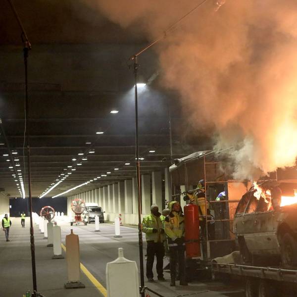 Flammen schlagen aus einem Schrottauto, das sich auf einem Anhänger befindet. Mehrere Personen beobachten das Geschehen.