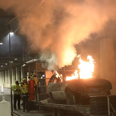 Flammen schlagen aus einem Schrottauto, das sich auf einem Anhänger befindet. Mehrere Personen beobachten das Geschehen.