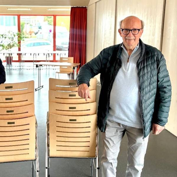 Zwei Männer stehen in einem großen Raum. Sie stützen jeweils einen Arm auf einen Stapel Stühle.