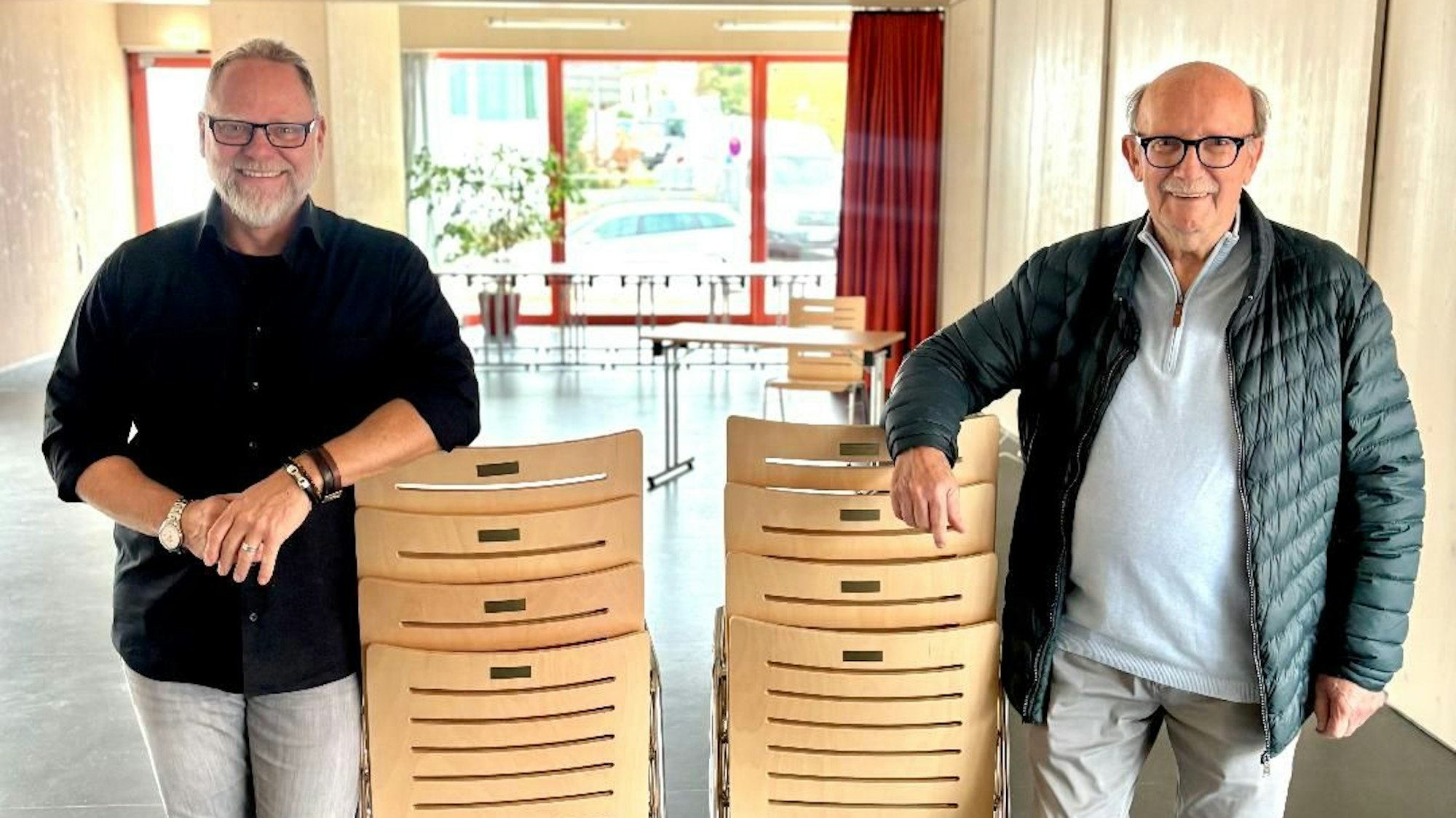 Zwei Männer stehen in einem großen Raum. Sie stützen jeweils einen Arm auf einen Stapel Stühle.