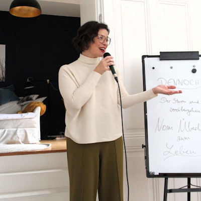 Eine Frau mit schwarzem Haar und Brille trägt roten Lippenstift und einen cremefarbenen Rollkragenpulli. Sie hält eine Mikro in der Hand.
