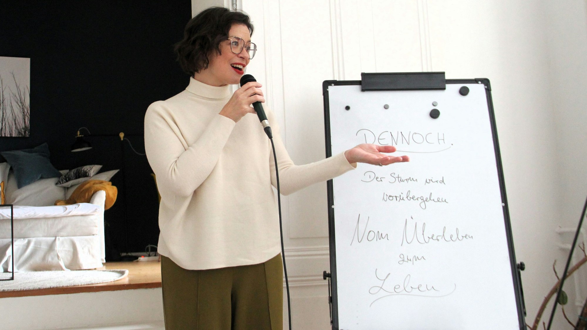 Eine Frau mit schwarzem Haar und Brille trägt roten Lippenstift und einen cremefarbenen Rollkragenpulli. Sie hält eine Mikro in der Hand.