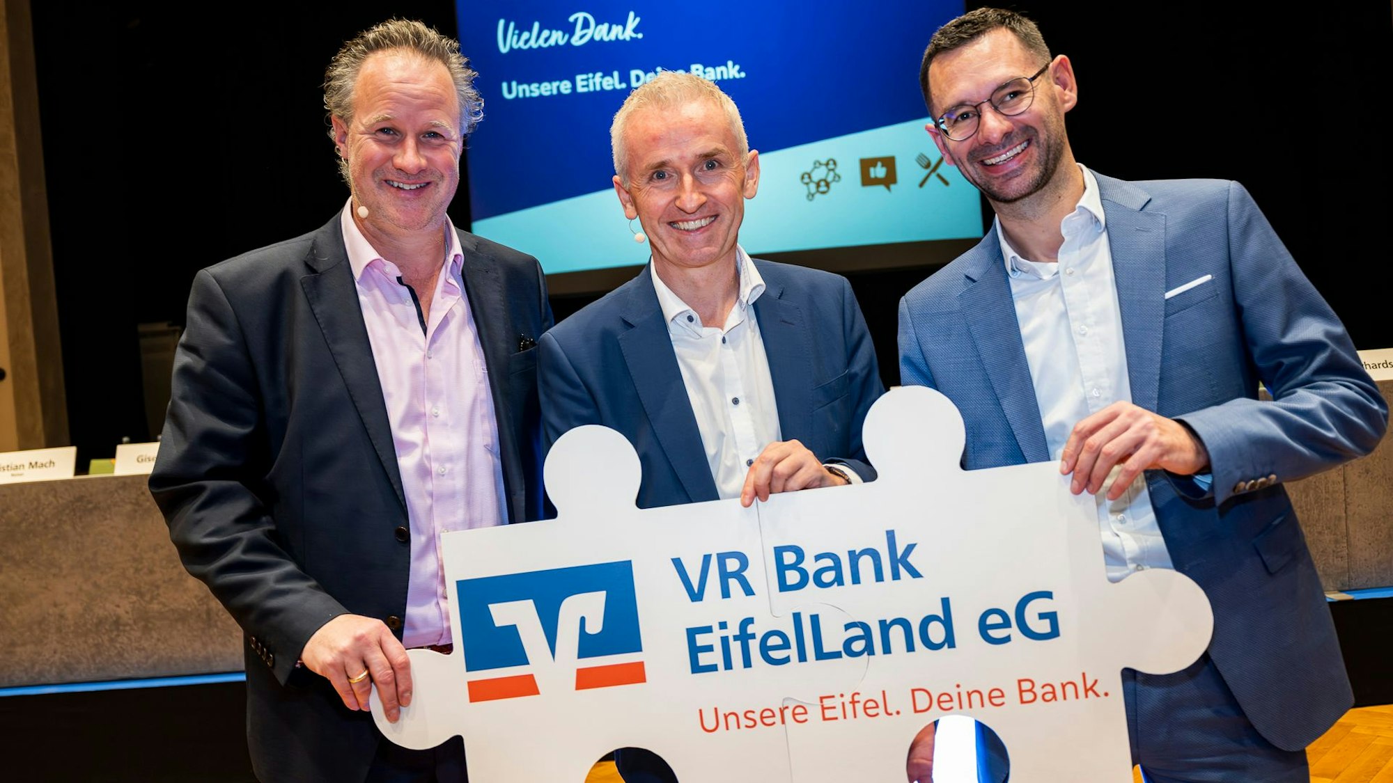 Drei Männer halten ein als Puzzleteile gestaltetes Plakat mit der Aufschrift VR Bank EifelLand eG.