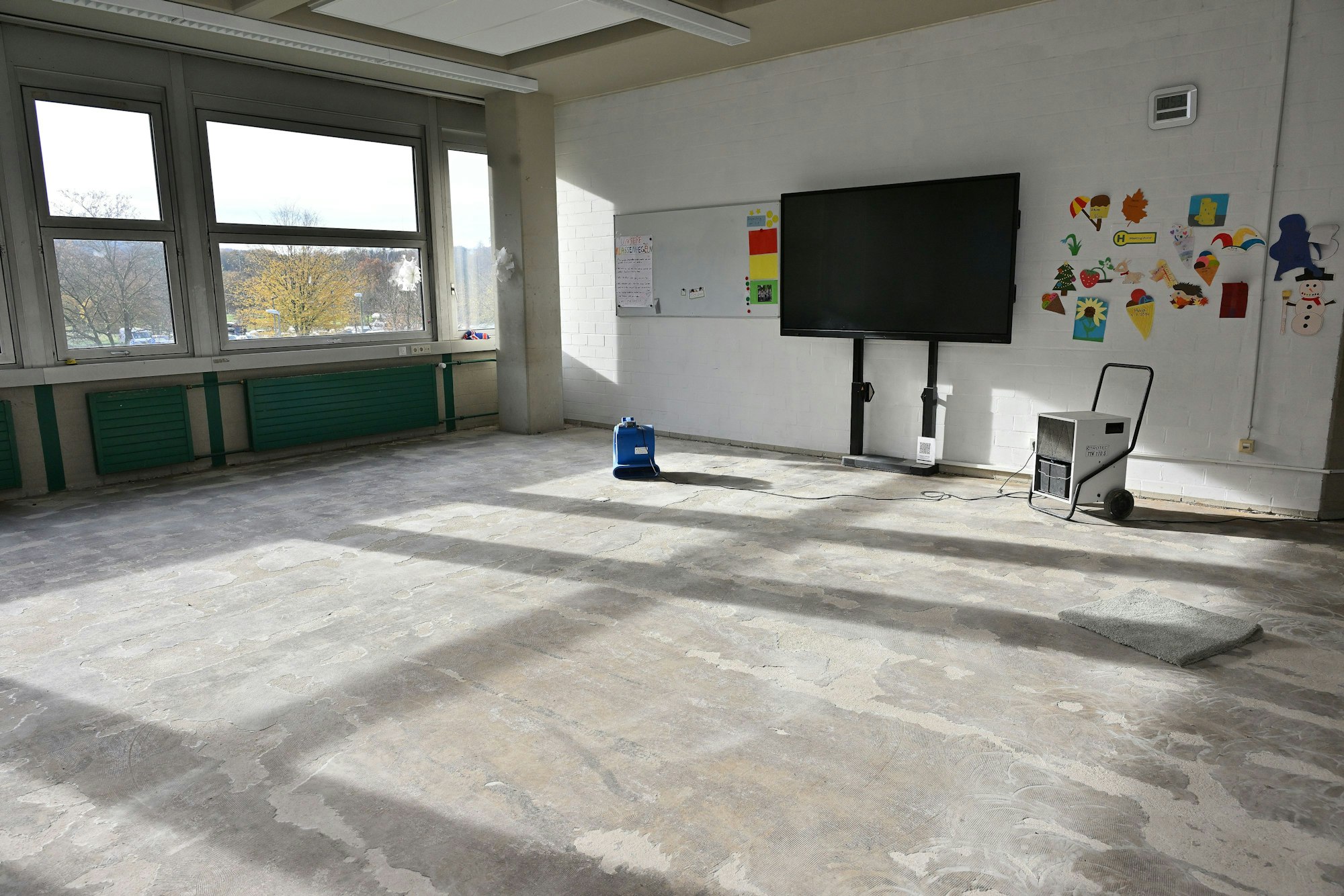 Ein leergeräumtes Klassenzimmer mit feuchtem Estrich und einem Bautrockner darin ist zu sehen.