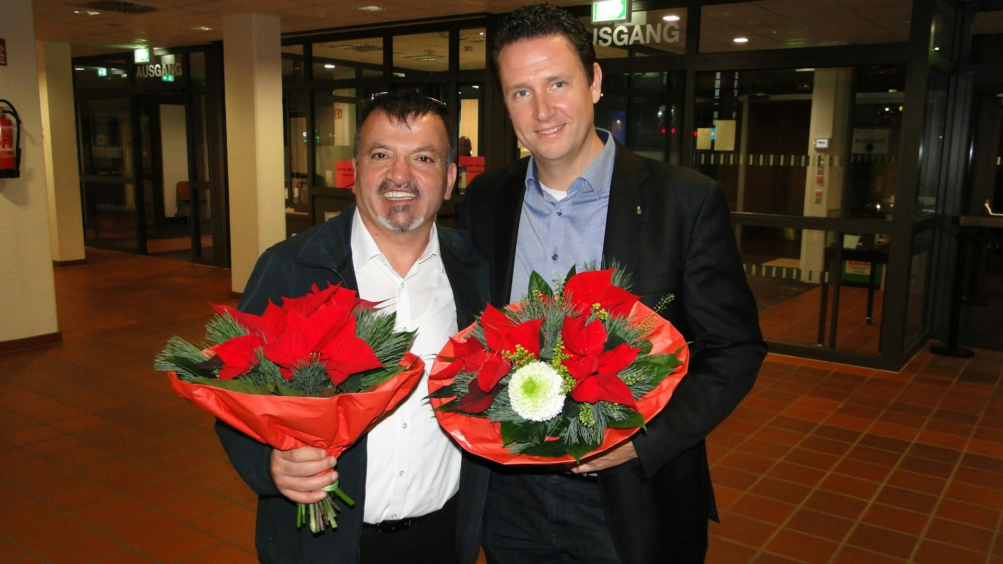 Zwei Männer stehen mit Blumensträußen in der Hand nebeneinander in einem Foyer.