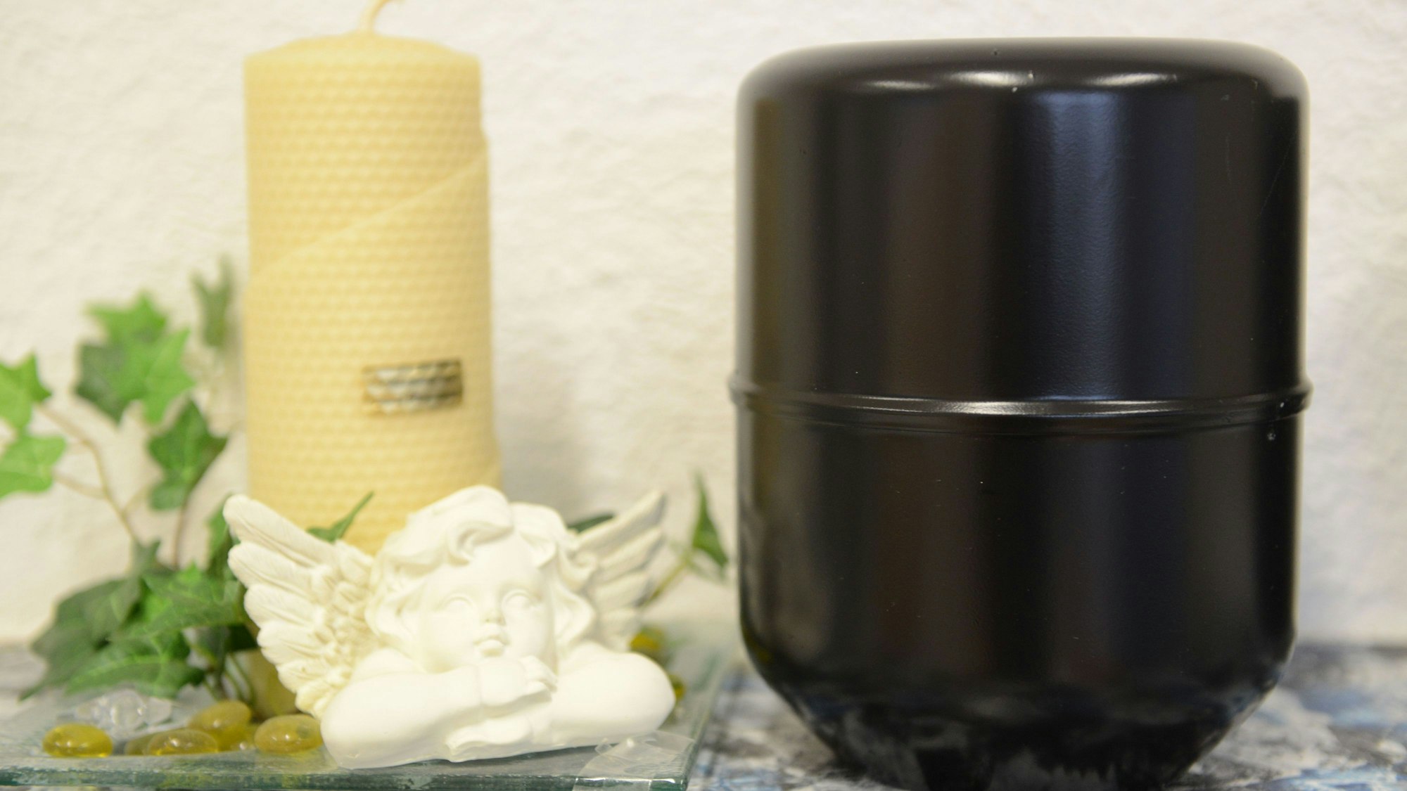 Eine Urne mit der Asche eines Verstorbenen steht bei einem Bestatter neben einer Kerze und einer Engelsfigur. (Symbolbild)