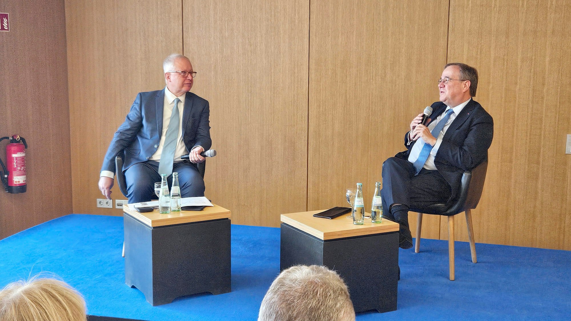 Tom Buhrow und Armin Laschet sitzen an zwei kleinen Tischen und haben jeder ein Mikro in der Hand.