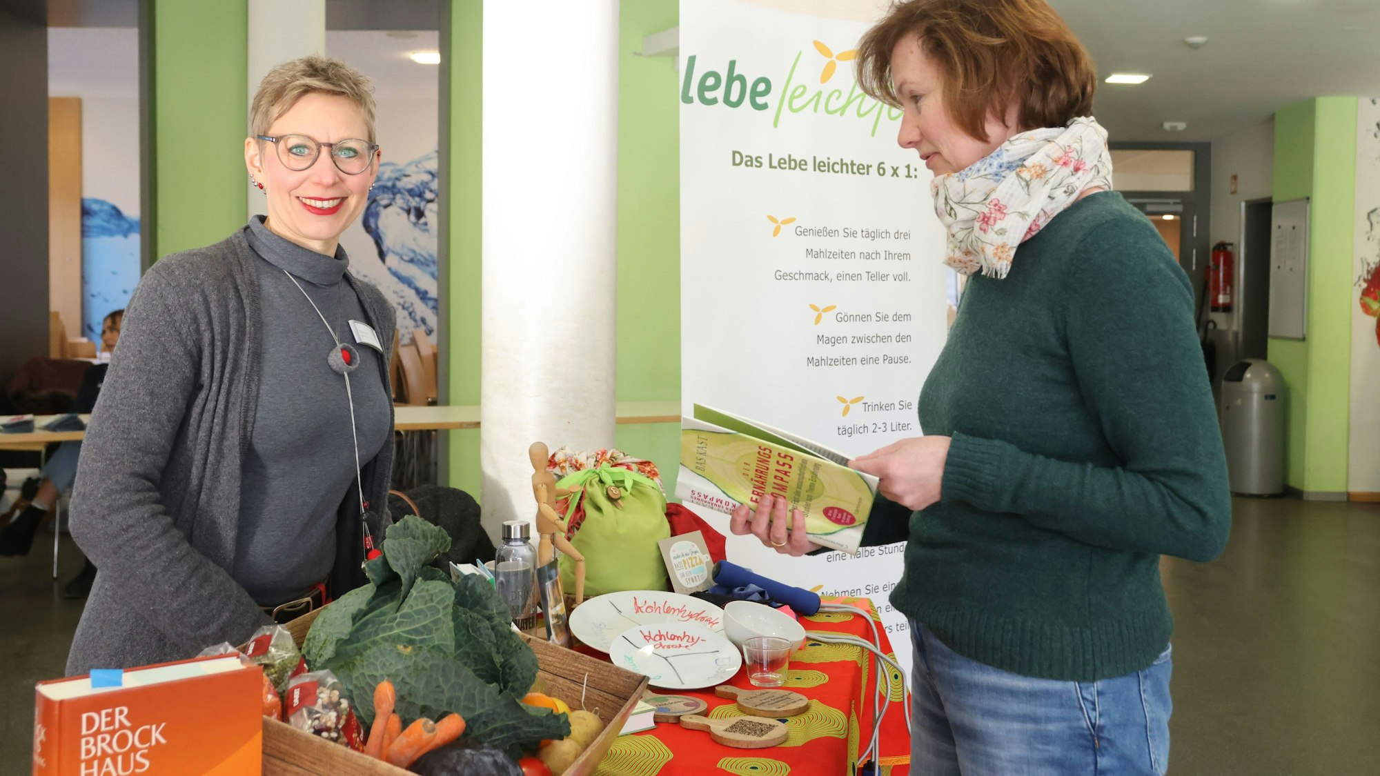 Das Foto zeigt eine Frau an einem Stand und eine zweite Frau, die in einer Broschüre blättert