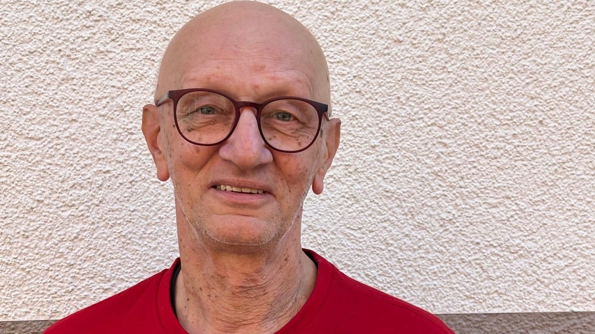 Auf dem Bild ist das Portrait eines Mannes in einem roten T-Shirt vor einer weißen Wand zu sehen.