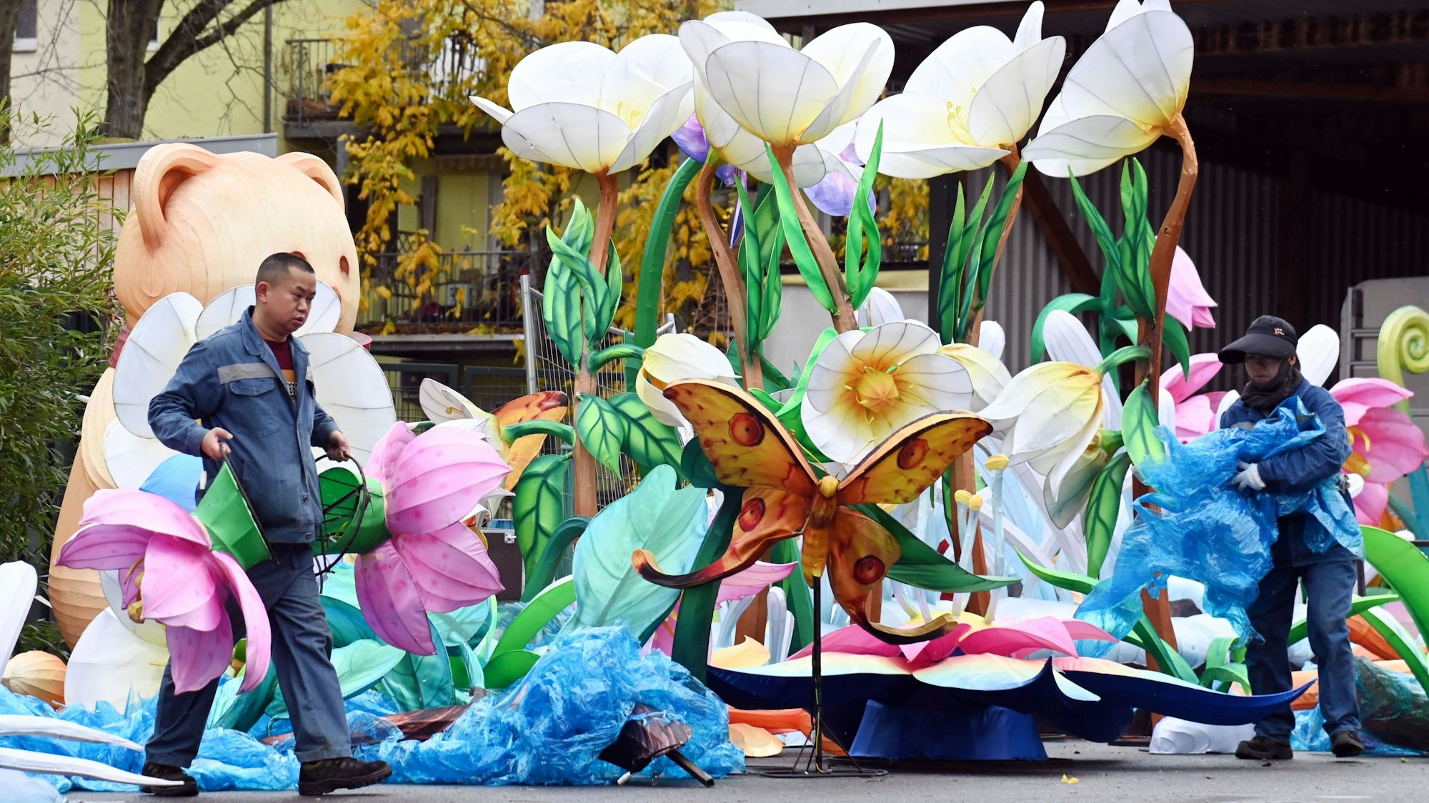 Ein Mann trägt zwei große, pinke Blumen. Ein anderer faltet blauen Stoff zusammen.