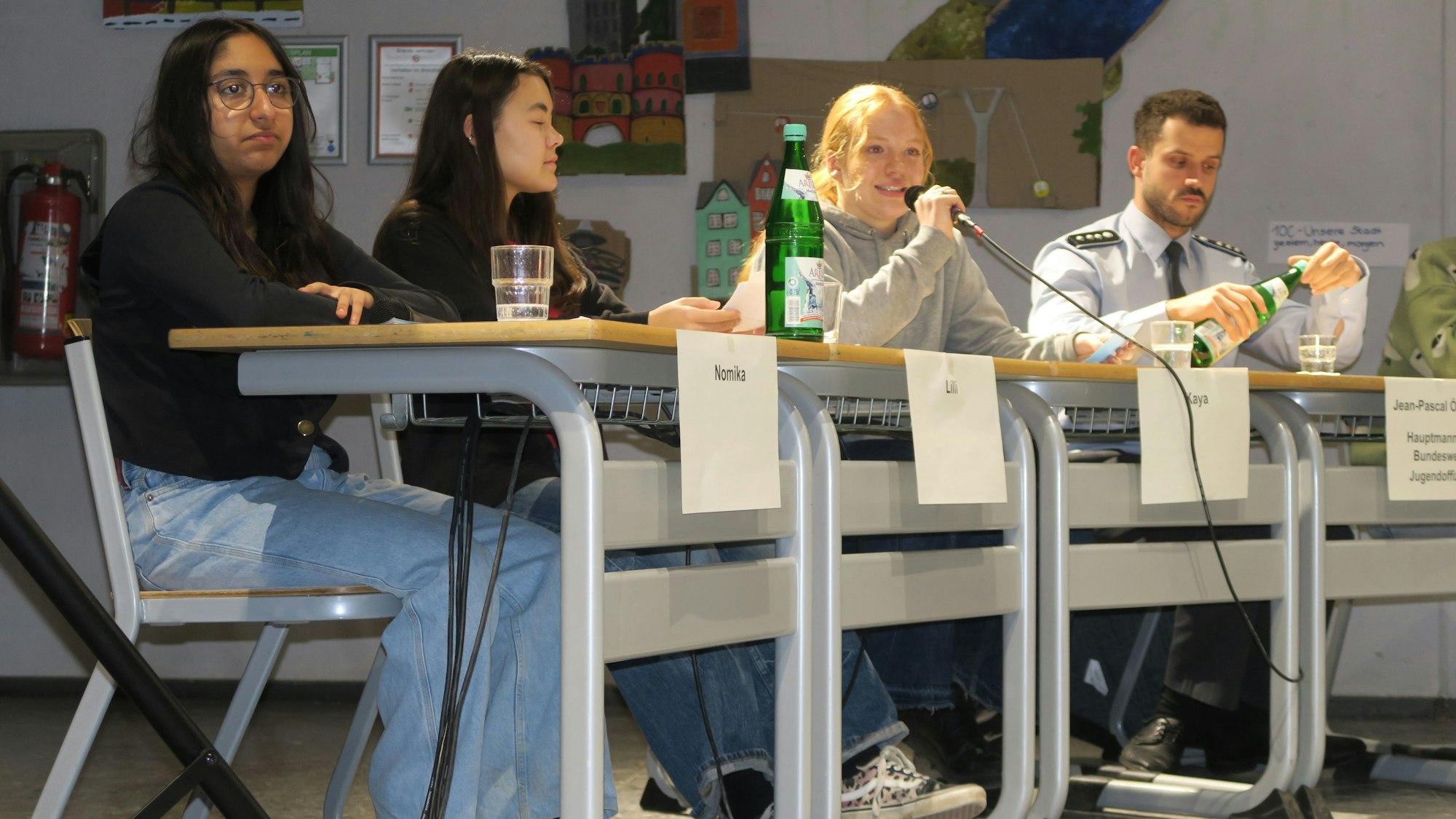 Drei Schülerinnen und ein Mann in Uniform sitzen nebeneinander.