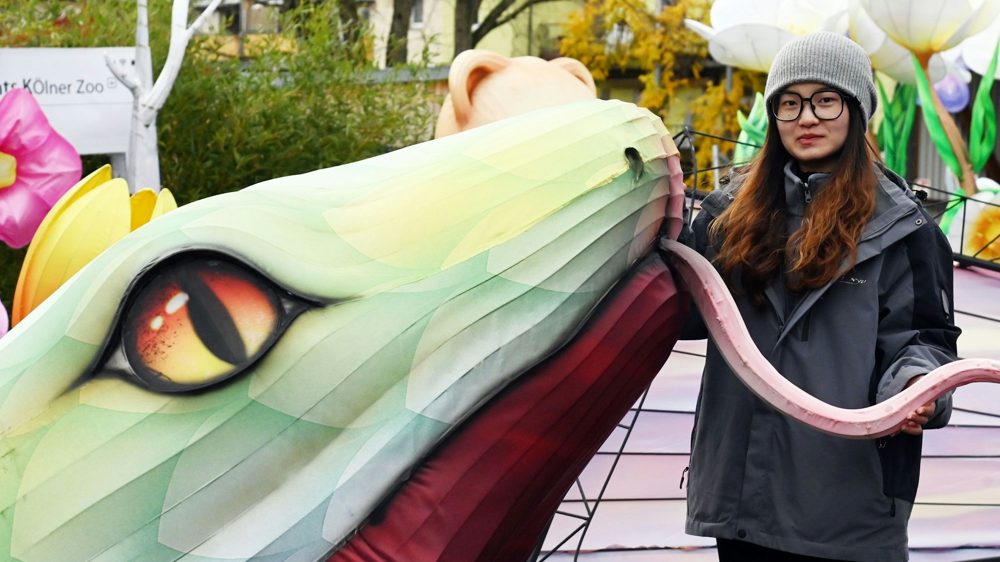 Eine Frau mit Brille und Mütze steht neben einem großen Schlangenkopf mit offenem Maul.