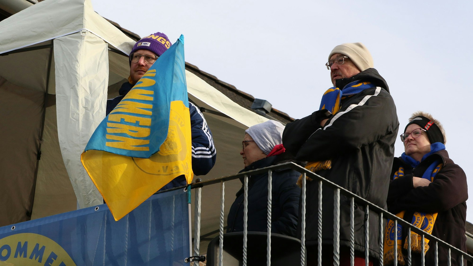 Hinter einer Kommern-Fahne steht Dirk Lamb mit einer lila Mütze. Neben ihm auf dem Balkon sind noch weitere Zuschauer.