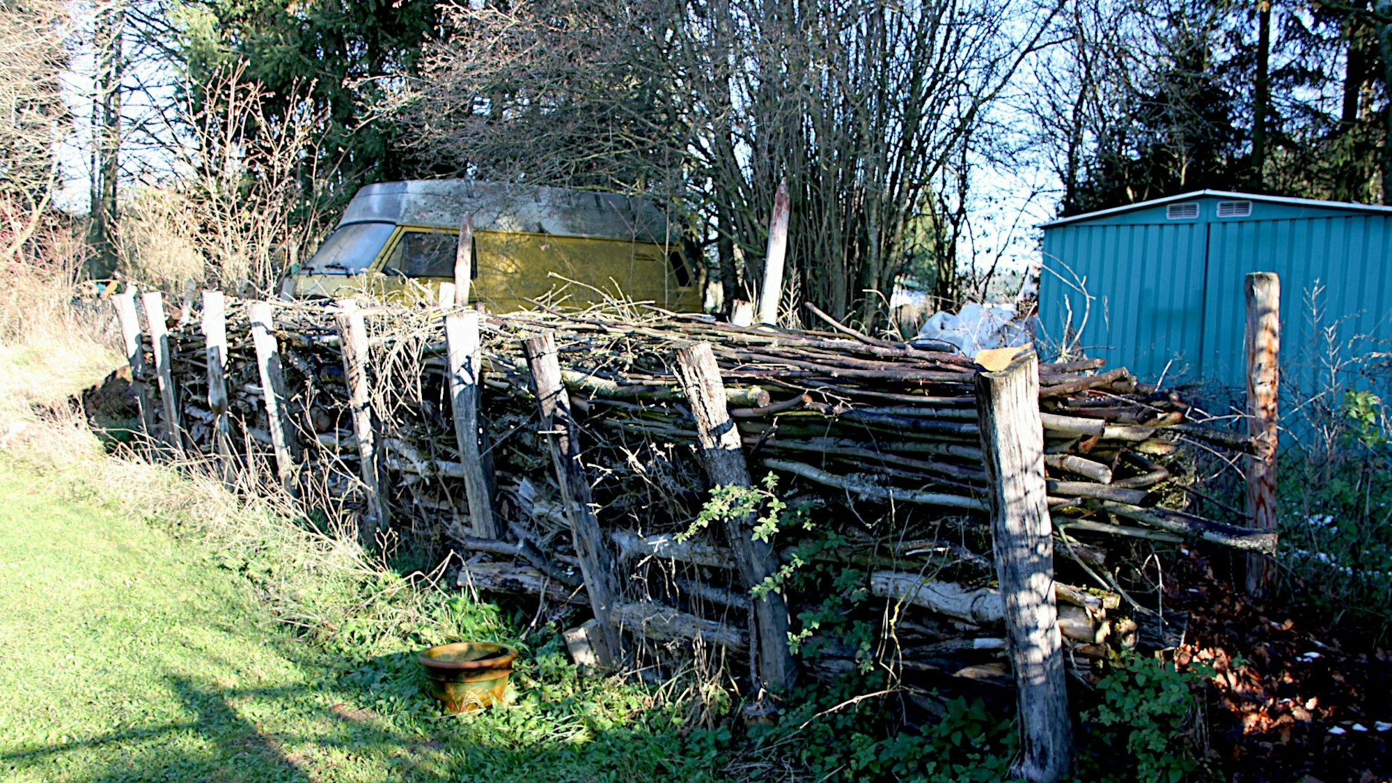 Eine Totholzhecke im Garten der Familie Böhm. Im Hintergrund sind ein blauer Gartenschuppen und ein gelber T3-VW-Bus mit weißem Hochdach zu sehen.