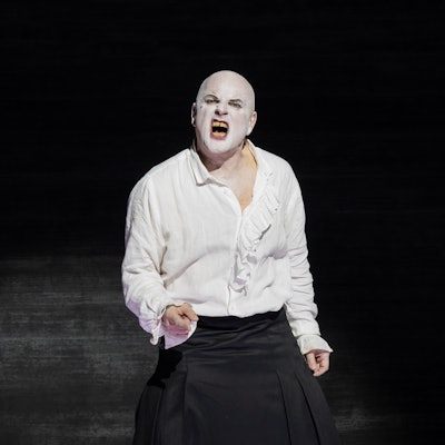 Zu sehen ist ein Schauspieler, der weiße Schminke auf dem Gesicht hat. Er verzerrt das Gesicht schmerzhaft im Gesang. Er trägt ein weißes Shirt und eine schwarze Hose, der Hintergrund ist schwarz.