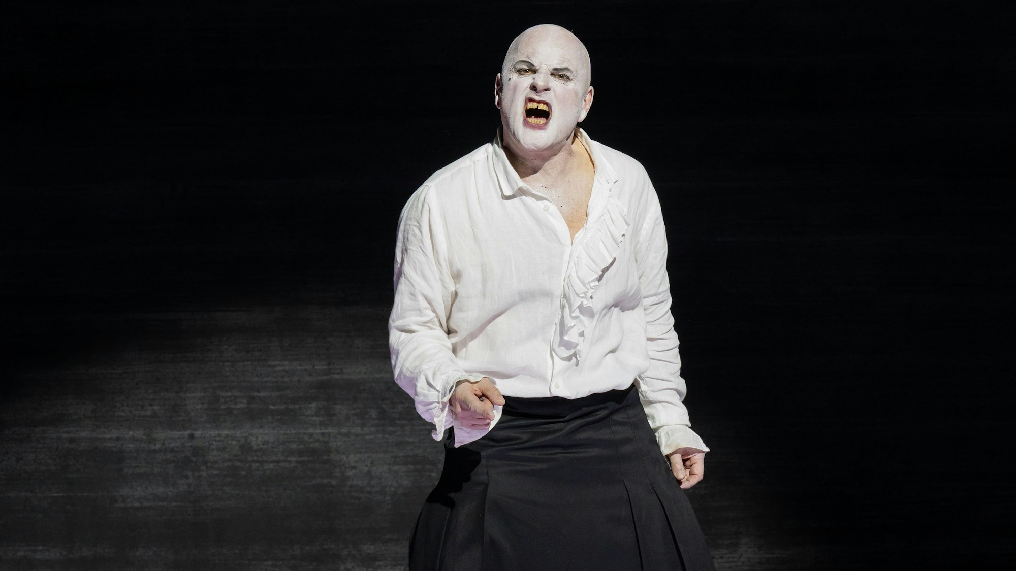Zu sehen ist ein Schauspieler, der weiße Schminke auf dem Gesicht hat. Er verzerrt das Gesicht schmerzhaft im Gesang. Er trägt ein weißes Shirt und eine schwarze Hose, der Hintergrund ist schwarz.