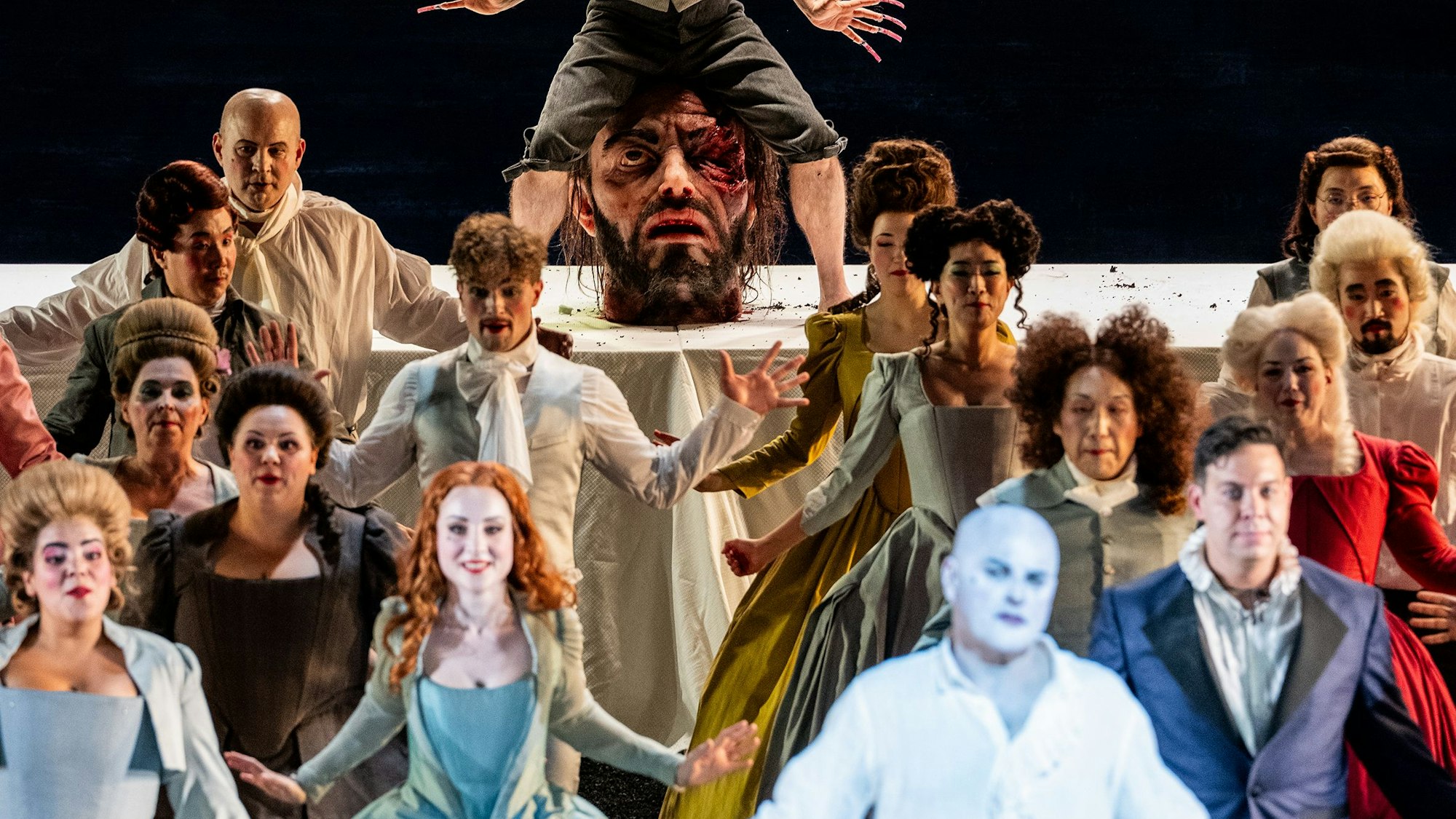 Vorne stehen die Darsteller in bunten Kostümen und breiten die Hände aus, im Hintergrund ein übergroßer Kopf auf der Bühne.