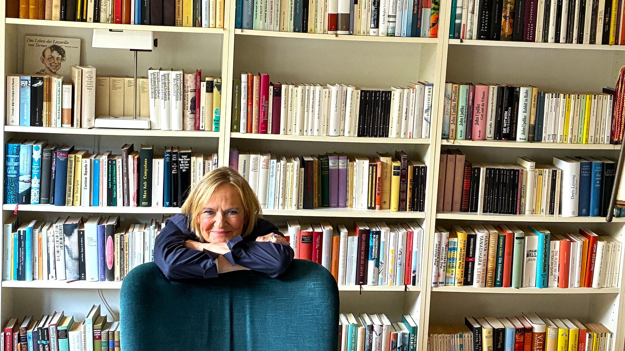 Eine Frau stützt die Arme auf einen Sessel, hinter ihr ist ein Bücherregal zu sehen.