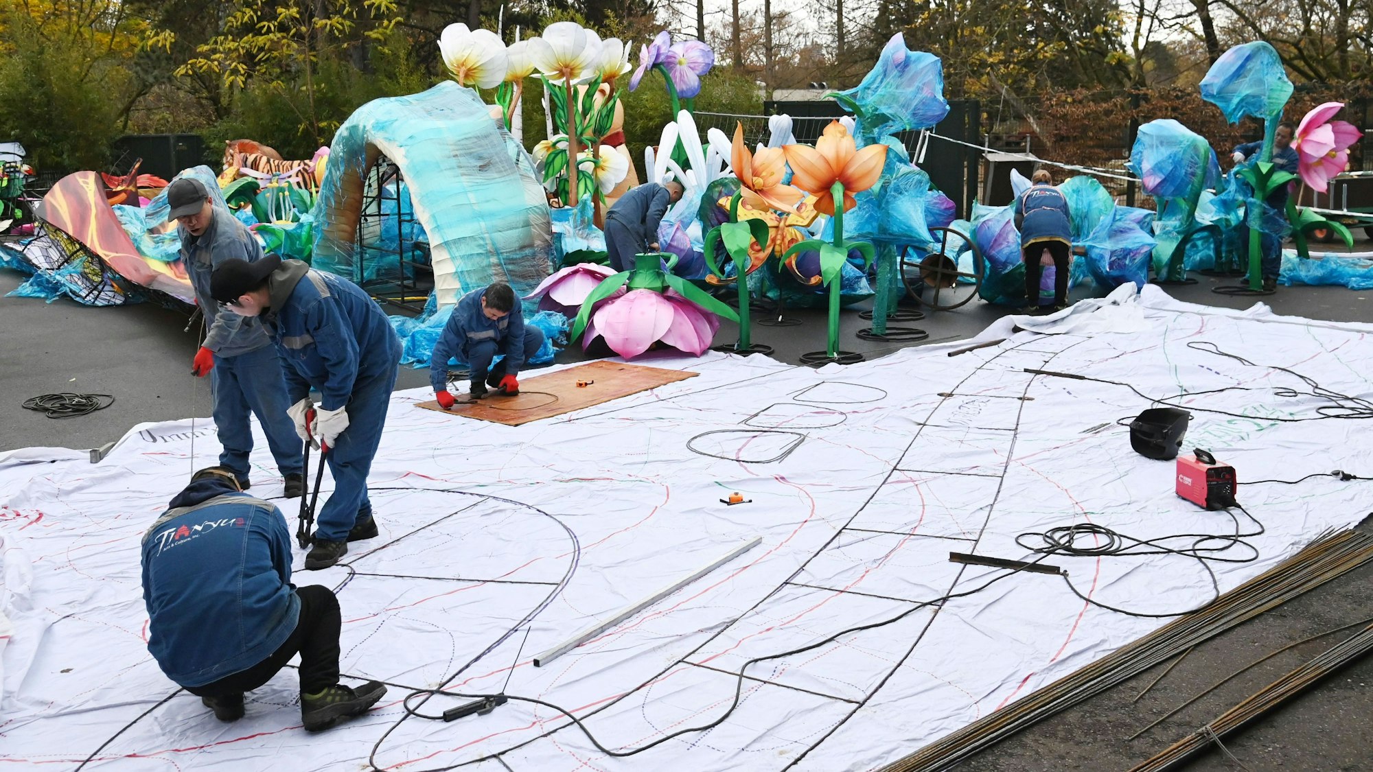 Auf dem Boden liegt eine weiße Plane, darauf stehen Männer und löten Eisenstangen zusammen. Im Hintergrund stehen große Blumen.