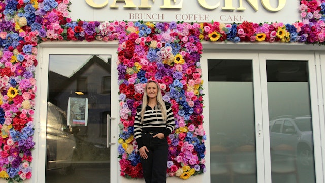 Eine Frau steht vor einer Fassade, die mit vielen Blumen geschmückt ist und den Schriftzug „Café Cino“ trägt.