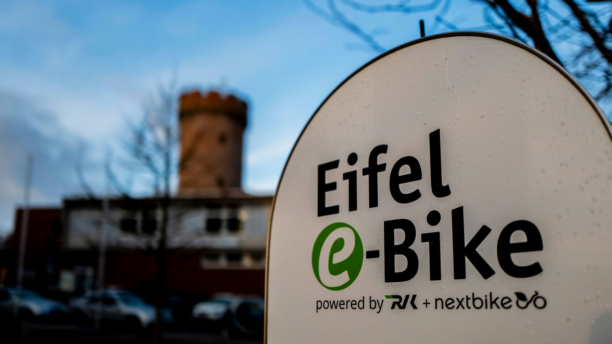 Das Bild zeigt das Hinweisschild auf die Eifel-E-Bike-Station. Im Hintergrund ist der Turm der Landesburg zu sehen.