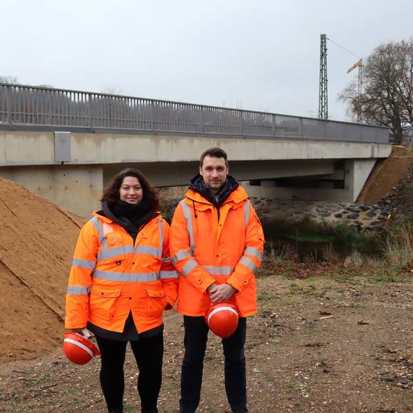Ein Mann und eine Frau in orangefarbener Jacke stehen vor einer Brücke. Beide halten einen Helm in ihren Händen.