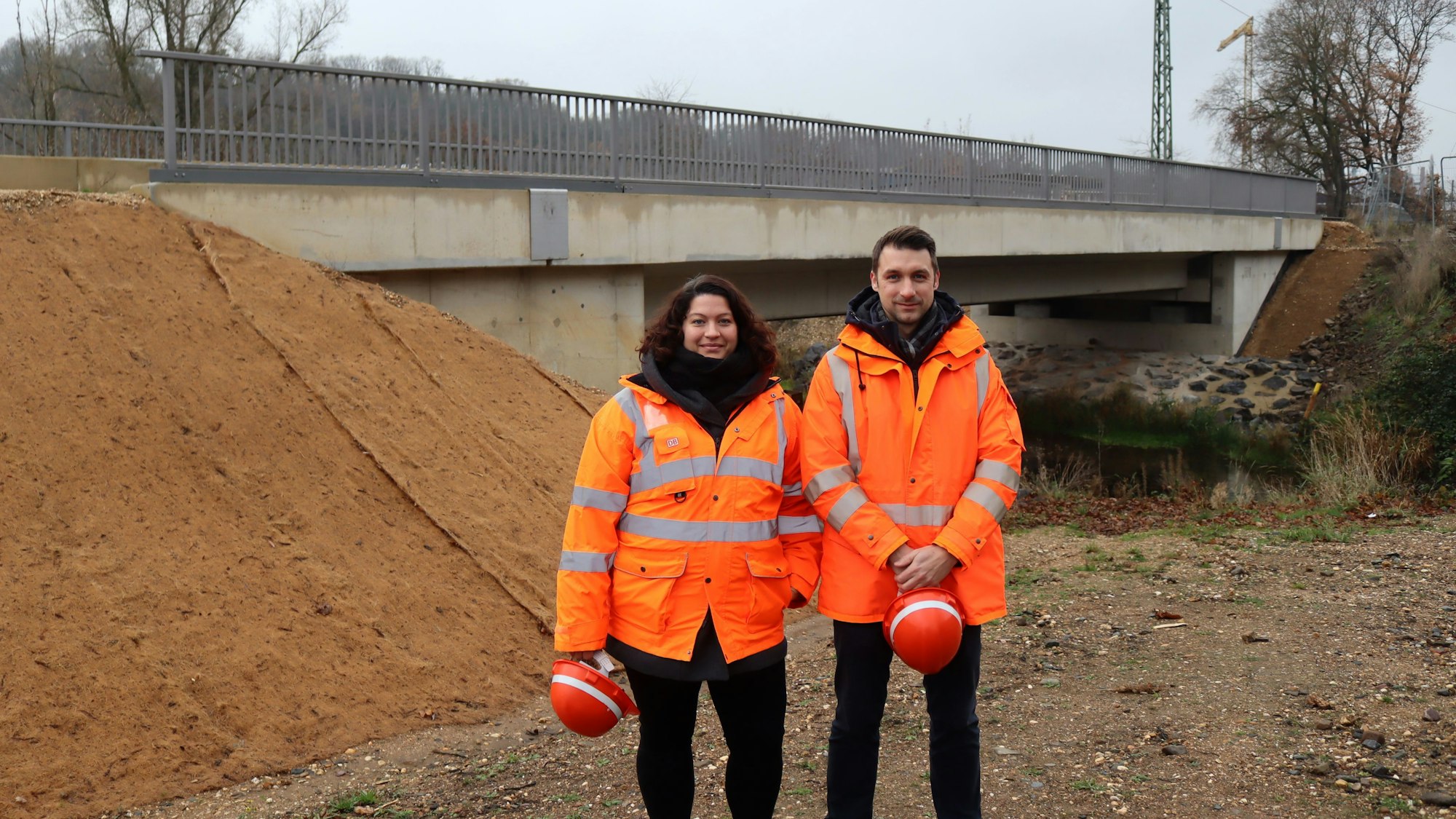 Ein Mann und eine Frau in orangefarbener Jacke stehen vor einer Brücke. Beide halten einen Helm in ihren Händen.
