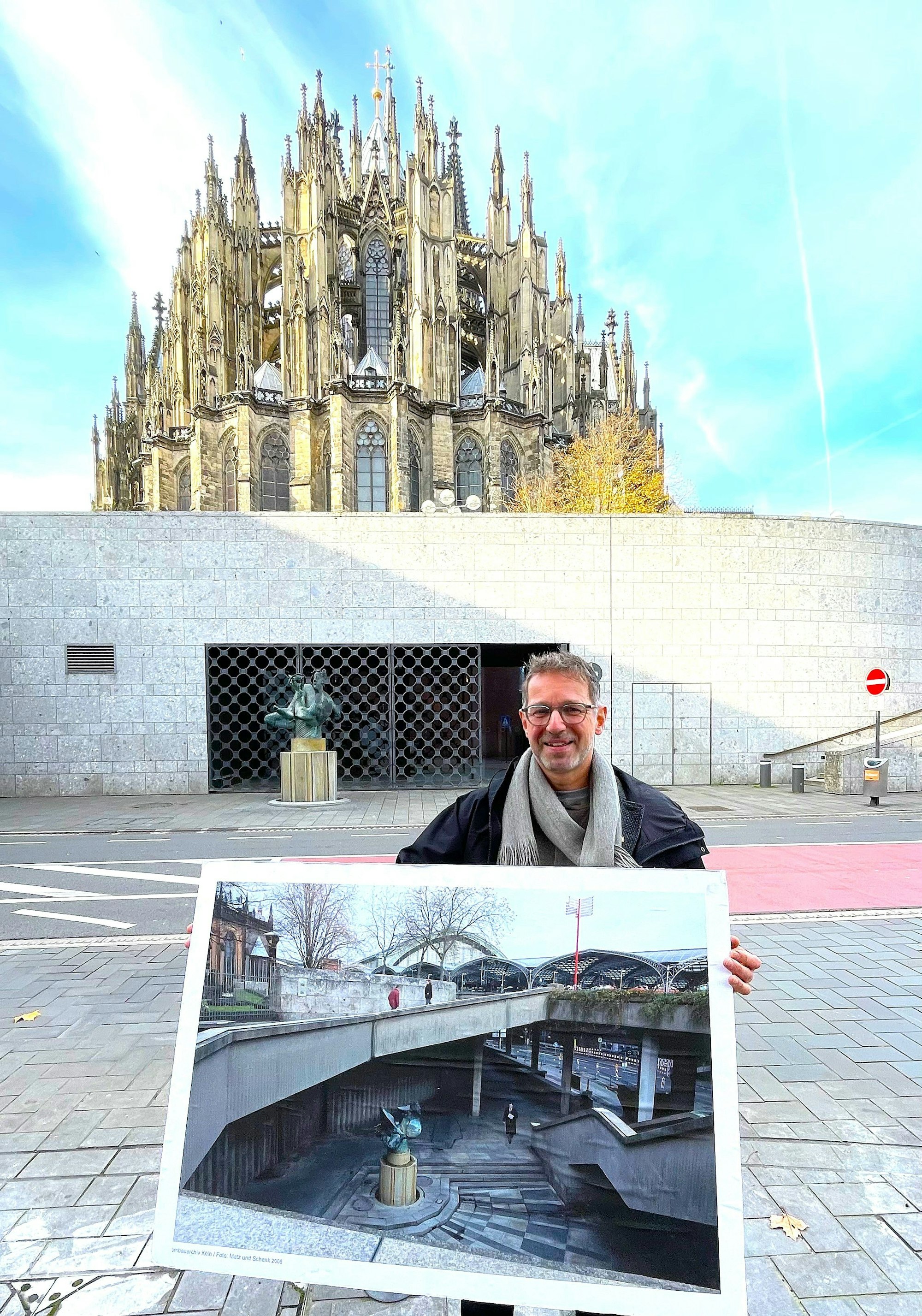 Ein Mann mit Brille steht vor einem vergitterten Eingang, dahinter ist das Baptisterium. In der Hand hält er ein Bild, das den Ort früher zeigt.