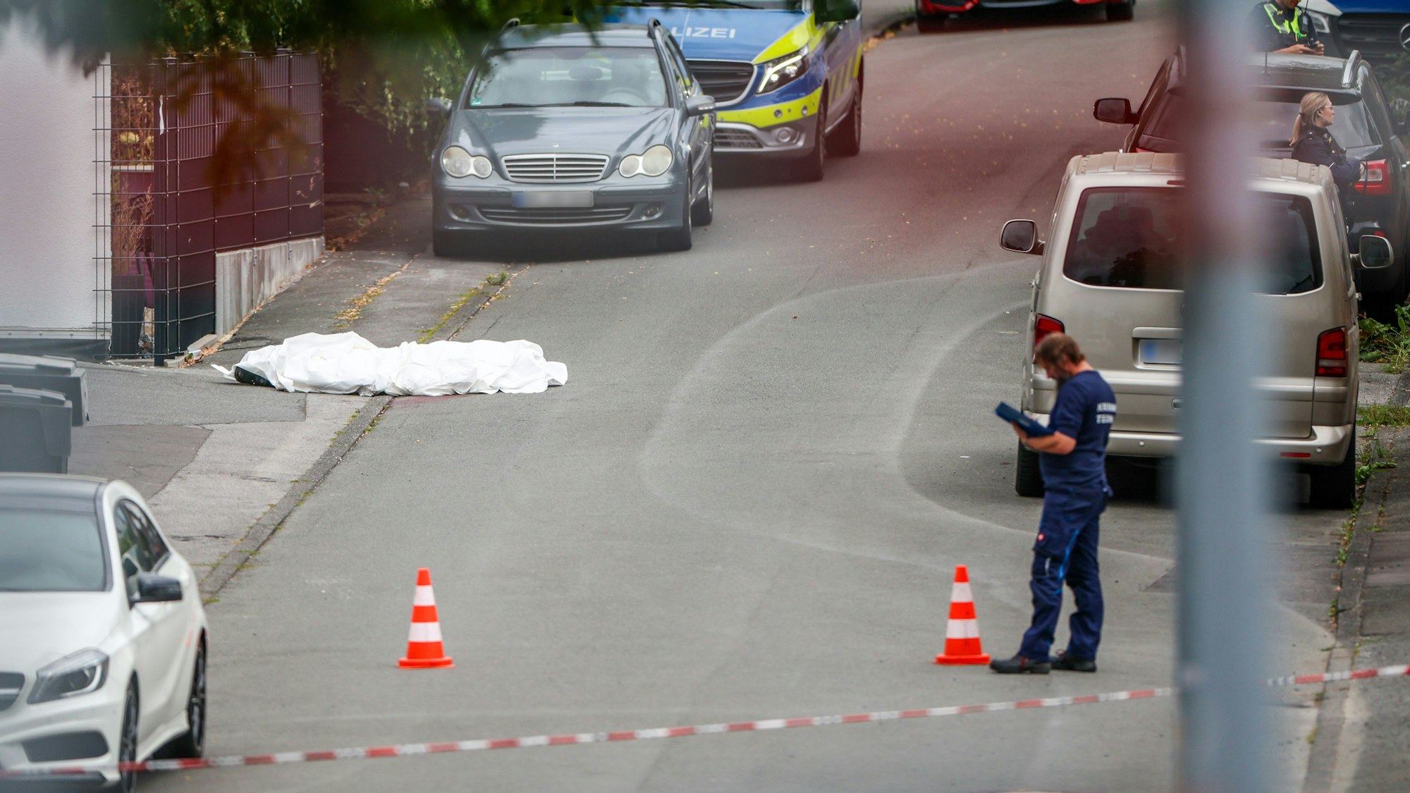 Ermittler der Spurensicherung der Polizei bei der Tatortarbeit nach Schüssen in Menden. Am Straßenrand auf der linken Seite liegt die abgedeckte Leiche einer erschossenen Person.