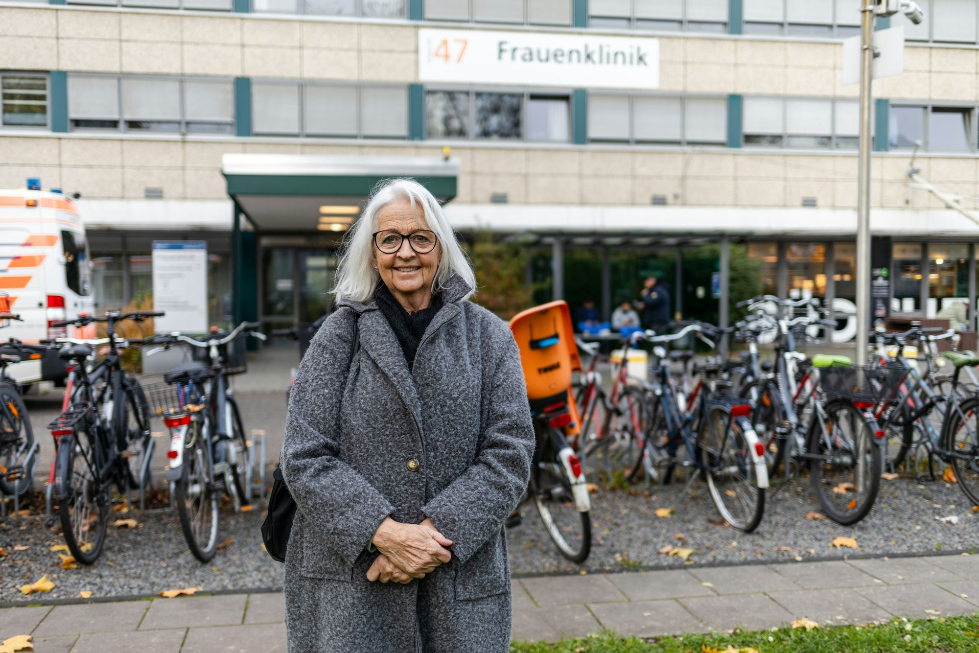 Eine Frau in grauem Mantel und mit grauen Haaren steht vor dem Eingang der Frauenklinik.