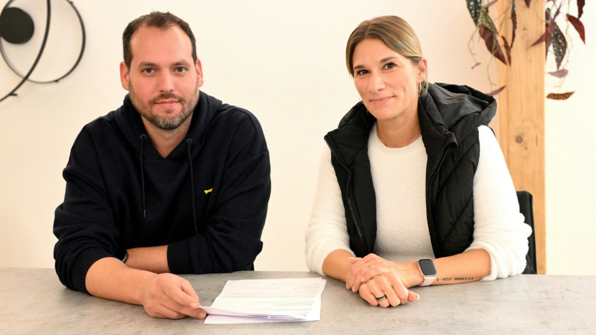 Ein Mann und eine Frau sitzen nebeneinander an einem Tisch mit Dokumenten vor sich liegen.