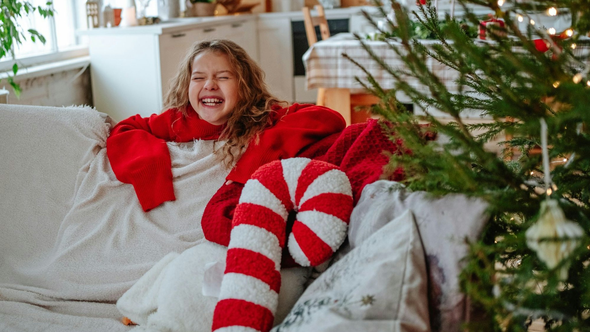 Ein Kind hinter einem Sofa, daneben steht ein Weihnachtsbaum, auf dem Sofa liegt ein Zuckerstangen-Kissen.