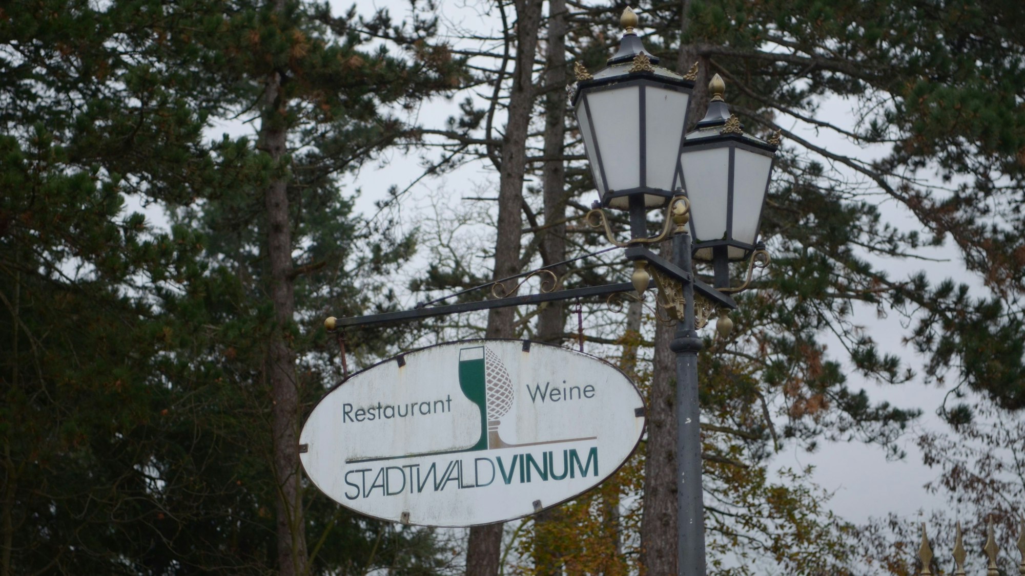 Das Schild ist an einer Doppellaterne befestigt, im Hintergrund stehen Bäume.