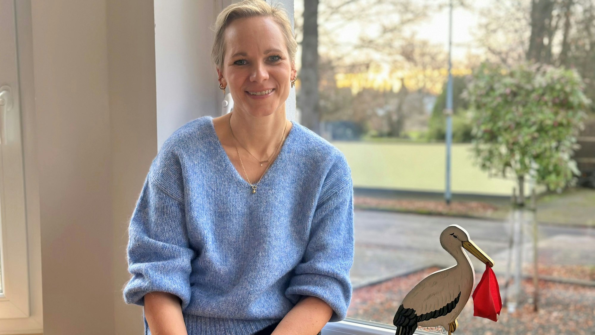 Eine Frau in blauem Pullover sitzt vor einem Fenster, neben ihr steht ein Storch aus Holz.