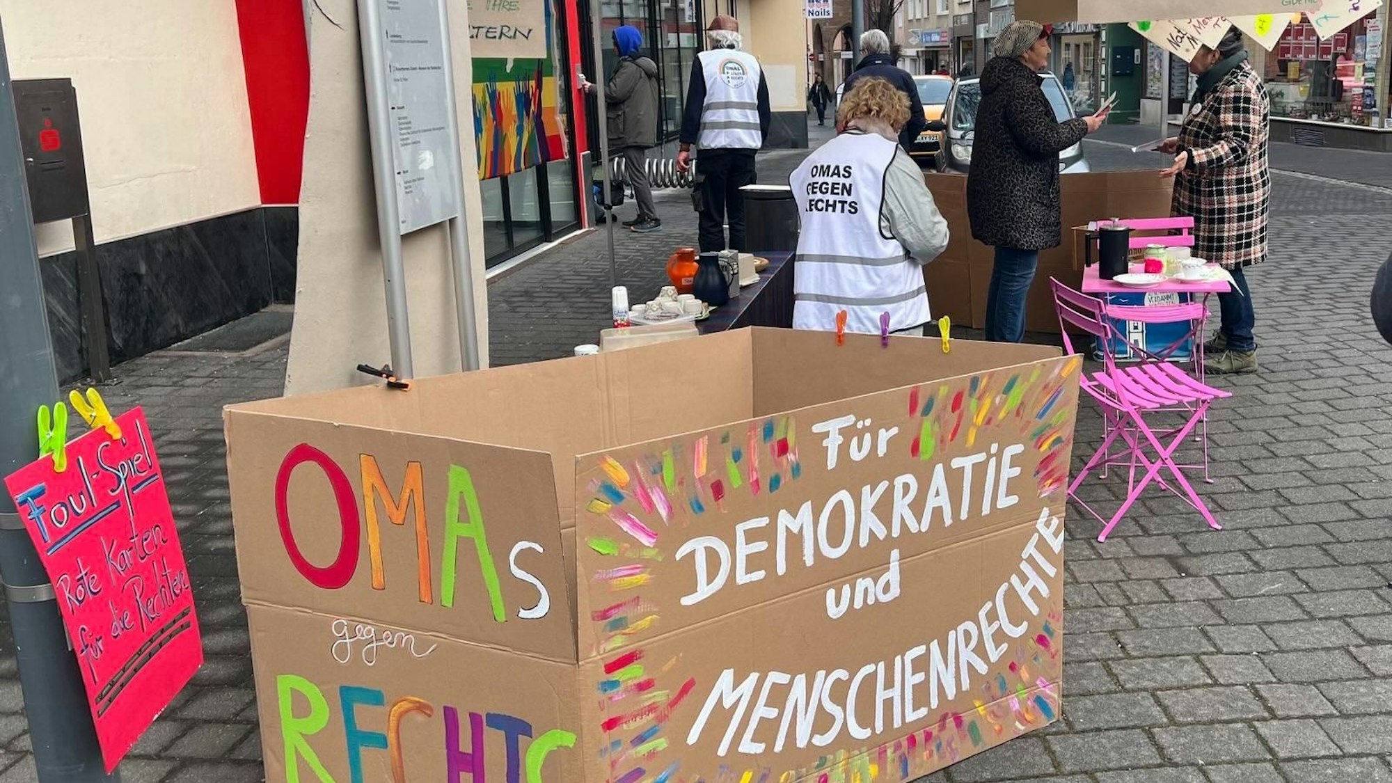 Ein Infostand der Zülpicher Omas gegen Rechts, im Vordergrund steht ein buntbemalter Karton mit dem Slogan Für Demokratie und Menschenrechte. Im Hitergrund sieht man einige der Aktivistinnen mit weißen Westen im Gespräch mit Passanten.