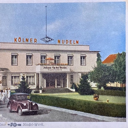 Frauen in weißen Kleidern und ein Auto stehen vor einem stattlichen Gebäude, auf dem der Schriftzug „Kölner JO Nudeln“ steht.