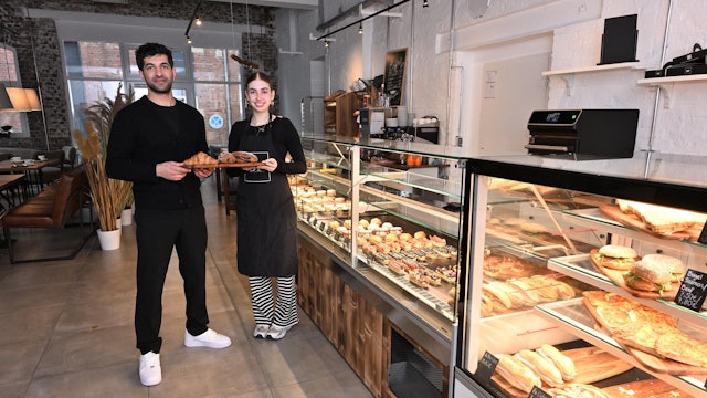 Die beiden stehen mit einem Tablett mit Gebäck vor der Theke.