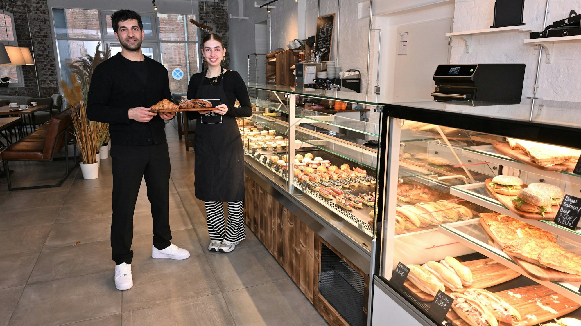 Die beiden stehen mit einem Tablett mit Gebäck vor der Theke.