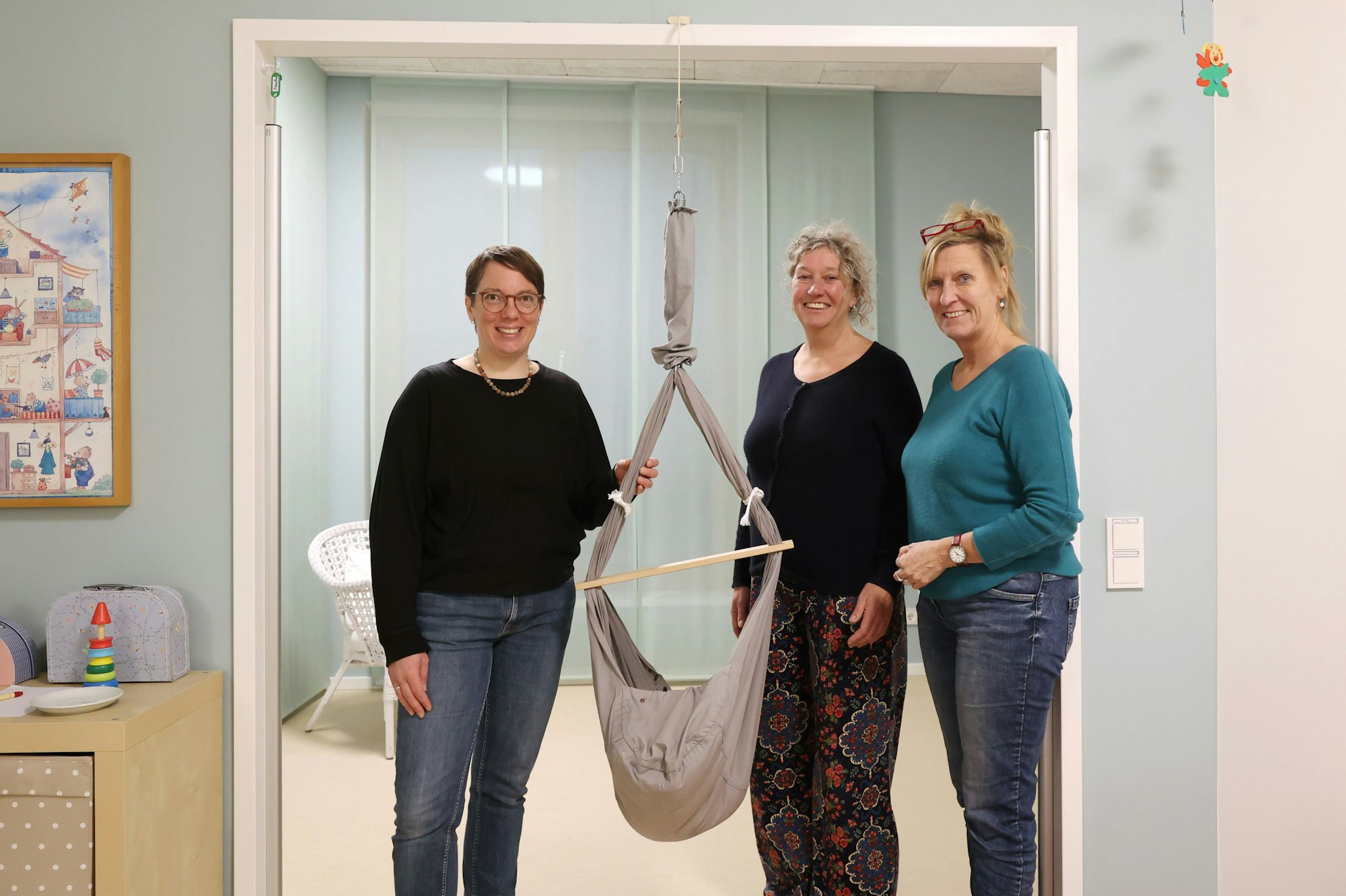 Die verantwortlichen SkF-Mitarbeiterinnen des Moses-Fensters Susanne Breyer, Corinna Sturm und Karin Horst stehen um eine Kinder-Wiege im freundlich eingerichteten Familien- und Kinderzimmer des Haus Adelheid.