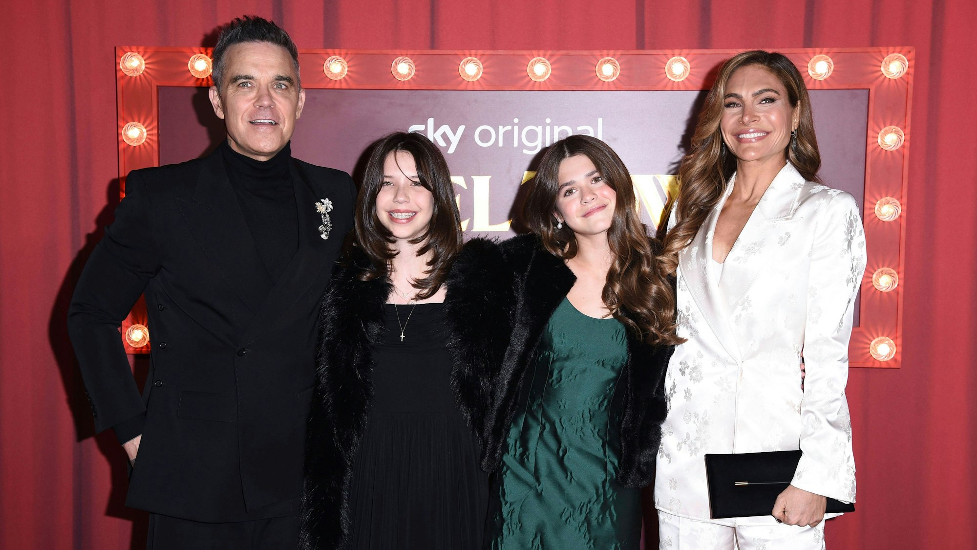Robbie Williams und Ayda Field mit ihren Töchtern lächelnd vor rotem Hintergrund.
