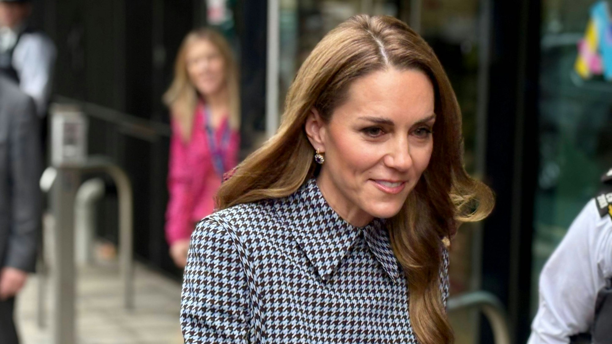 Kate Middleton in einem schwarz-hellblauen Hahnentrittmuster-Kleid mit unscharfen Personen im Hintergrund.