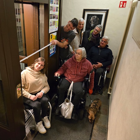 Eine Gruppe von Menschen, einige im Rollstuhl, einer misst die Breite eines Fahrstuhlzugangs.